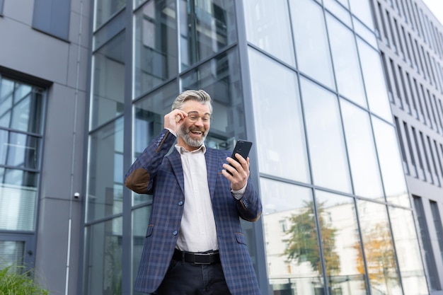 Un homme senior aux cheveux gris heureux dans un costume et des lunettes se tient à l'extérieur d'un bureau un gratte-ciel joyeux