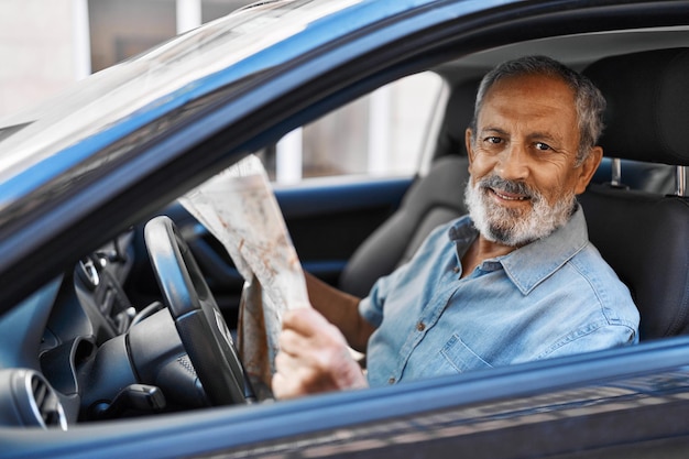 Homme senior aux cheveux gris tenant un plan de la ville assis sur une voiture dans la rue