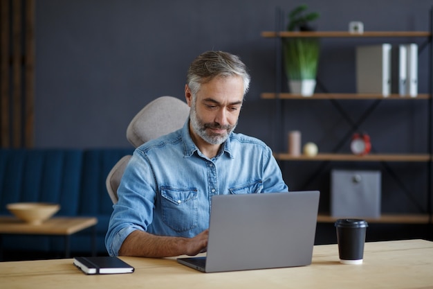 Homme senior aux cheveux gris travaillant au bureau à domicile avec ordinateur portable.