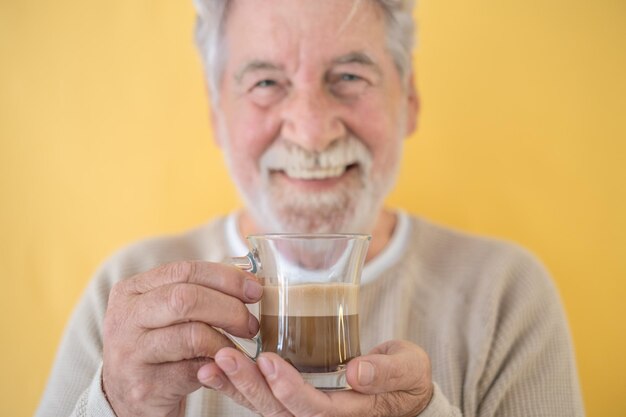 Homme senior barbu attrayant défocalisé tenant un verre de café et de lait, cappuccino, debout sur fond jaune