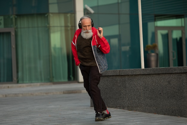 Un homme senior barbu branché, écouter de la musique en plein air. Homme mature de mode.