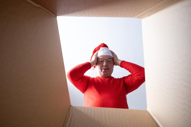 Photo homme senior en casquette de noël déballant, ouvrant une boîte en carton et regardant à l'intérieur.