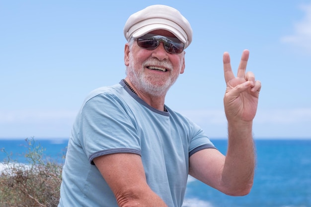 Homme senior caucasien souriant avec un chapeau assis sur la plage regardant la caméra gesticulant le signe de la victoire tout en profitant des vacances ou de la retraite Horizon sur l'espace de copie de l'eau