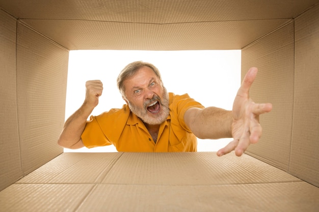 Homme senior en colère ouvrant le plus gros colis postal isolé sur blanc. Triste modèle masculin sur le dessus de la boîte en carton regardant à l'intérieur.