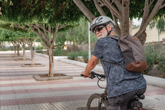 Homme senior cycliste actif souriant dans un parc urbain portant un casque et un sac à dos en cours d'exécution avec un vélo électrique Concept de mode de vie sain et de mobilité durable