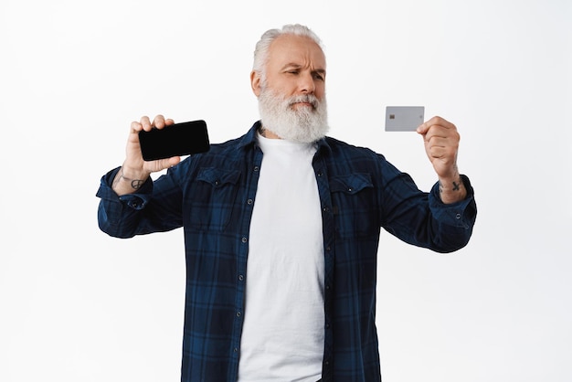 Homme senior douteux et sceptique montrant l'écran horizontal du smartphone et regardant avec incrédulité la carte de crédit ayant des doutes n'étant pas sûr debout sur fond blanc