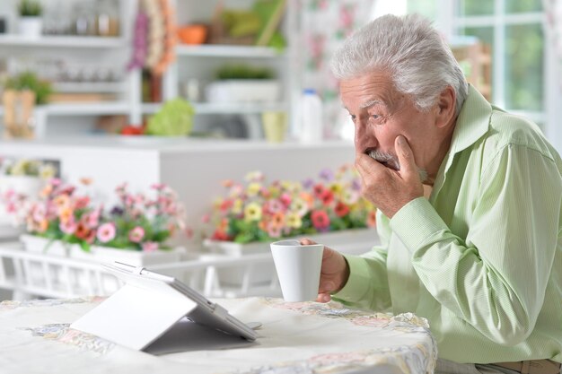 Homme senior émotionnel regardant une tablette numérique tout en buvant du thé