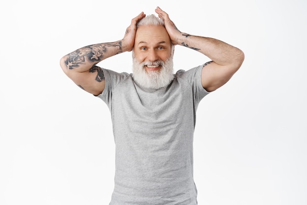 Un homme senior excité et heureux avec des tatouages entend des nouvelles surprises incroyables tenant les mains sur la tête et souriant étonné d'être impressionné par un grand événement debout en t-shirt gris sur fond blanc