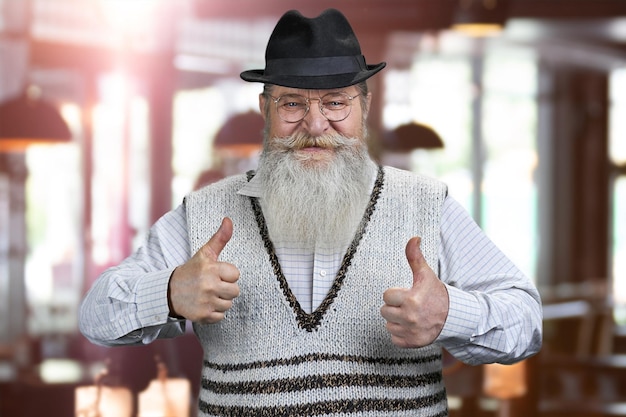 Homme senior joyeux avec barbe regardant la caméra et gesticulant les pouces vers le haut.