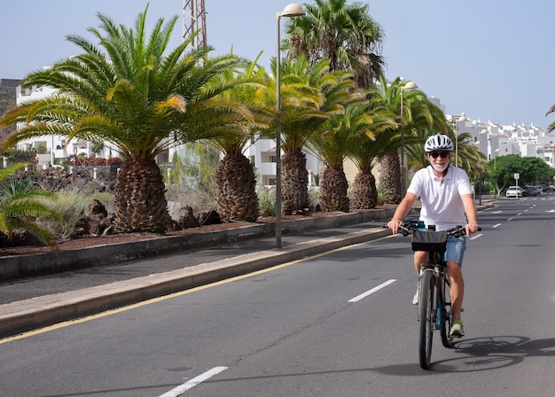 Homme senior joyeux avec casque appréciant l'activité sportive en cours d'exécution avec son vélo électrique dans une rue ensoleillée palmiers sur fond concept de mobilité durable