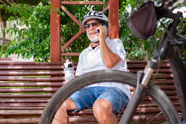 Homme senior joyeux avec casque et vélo assis dans un parc public parlant sur un téléphone portable homme âgé tenant une bouteille d'eau
