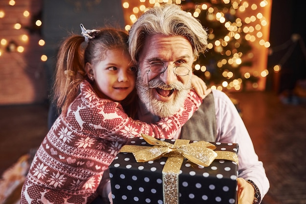 Homme senior à la mode gai aux cheveux gris et à la barbe assis avec une petite fille dans une salle de noël décorée.