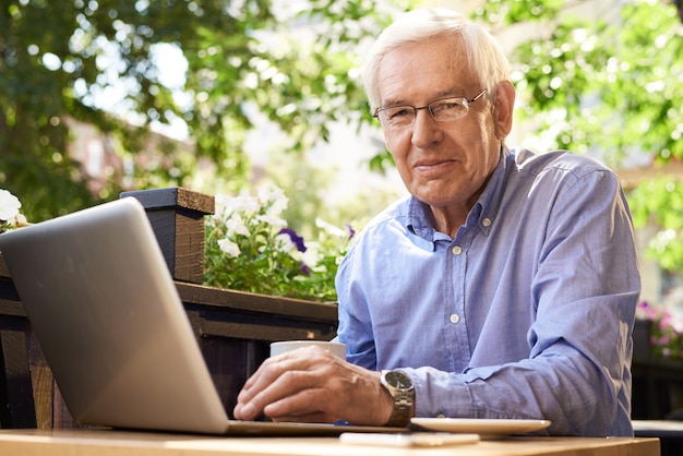 Homme senior moderne à l'aide d'un ordinateur portable dans un café en plein air