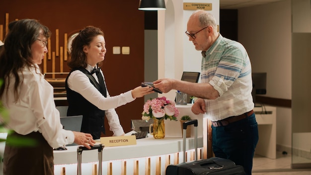 Photo homme senior payant pour une chambre d'hôtel avec des services tout compris, utilisant une carte pour les transactions électroniques au terminal de point de vente. couple à l'âge de la retraite voyageant pour ses loisirs, se relaxant dans un complexe cinq étoiles.