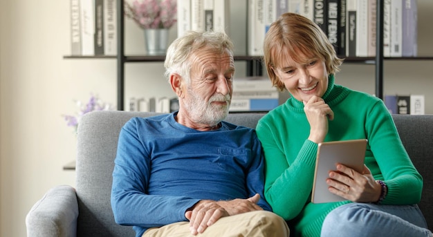 Homme senior positif et femme mature avec tablette souriant et regardant la caméra assis sur un canapé dans le salon ensemble