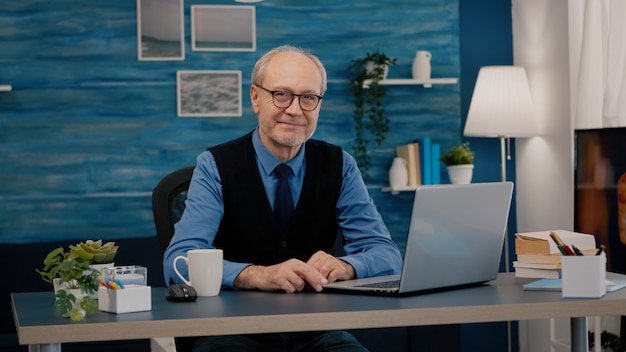 Homme senior regardant la caméra souriant assis sur le lieu de travail au bureau travaillant à domicile pendant que la vieille femme regarde la télévision en arrière-plan. Homme d'affaires à la retraite préparé pour une réunion en ligne à l'aide de gadgets modernes