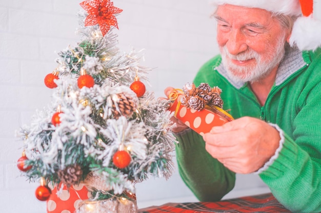Homme senior à la retraite souriant en bonnet de noel et vêtements chauds décorant l'arbre de noël à la maison. Vieux père Noël souriant tout en regardant l'arbre de Noël décoré. Homme âgé profitant des vacances de Noël