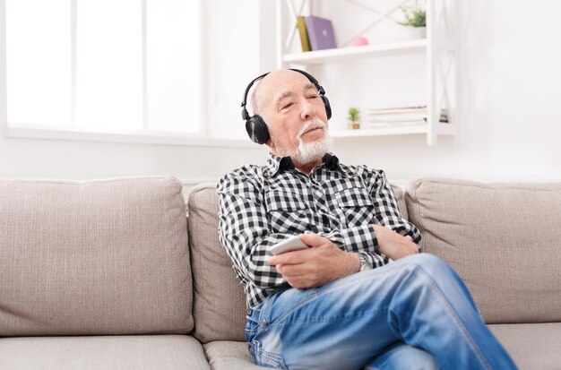 Homme senior rêveur écoutant de la musique sur un smartphone avec un casque, assis sur un canapé à la maison, espace pour copie