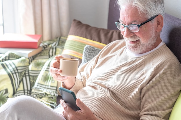 Homme senior séduisant, cheveux blancs, assis sur un canapé à la maison, buvant un café en regardant un téléphone portable
