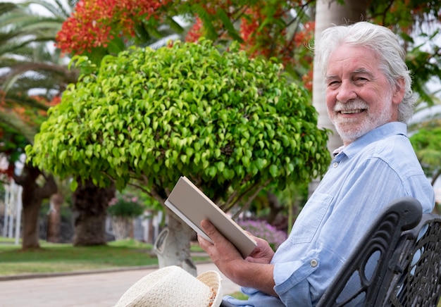 Homme senior séduisant avec des sourires de barbe blanche assis dans le parc en lisant un livre