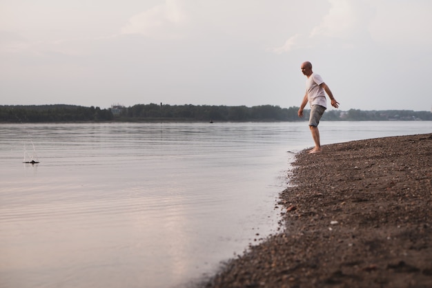 Homme senior solitaire marchant pieds nus au bord de la rivière et jetant des cailloux dans l'eau