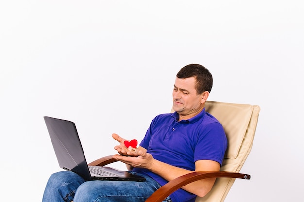 Un homme senior sourd satisfait tenant une aide auditive et un cœur rouge devant une caméra d'ordinateur portable