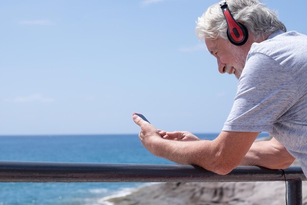 Homme senior souriant adulte à l'extérieur en mer profitant d'une journée ensoleillée à l'aide d'un téléphone portable portant des écouteurs