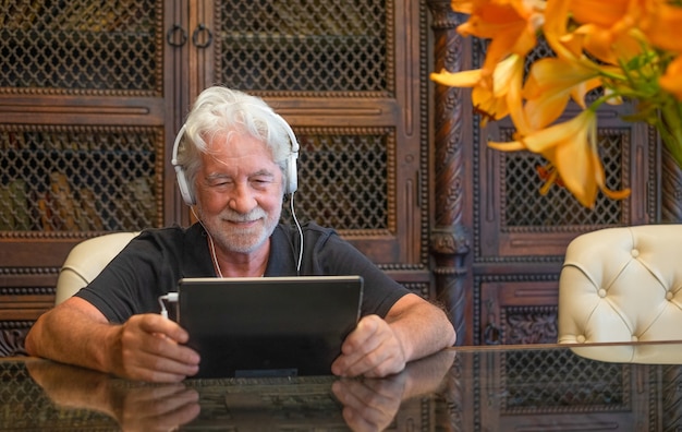 Homme senior souriant à l'aide d'un appareil électronique avec un casque. Assis à l'intérieur à une table en bois
