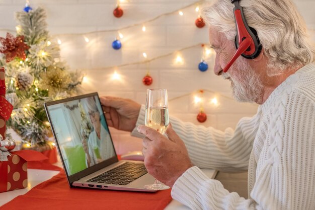 Homme senior souriant en appel vidéo avec ordinateur portable célébrant Noël avec un verre de vin mousseux. Bonne année et joyeux Noël