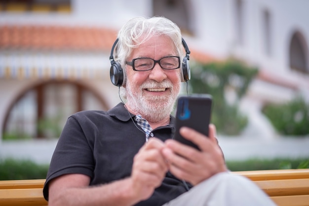 Homme senior souriant en appel vidéo avec smartphone et casque assis en plein air sur un banc