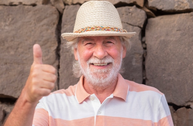 Homme senior souriant aux cheveux blancs et chapeau de paille faisant signe ok avec les mains debout en plein air