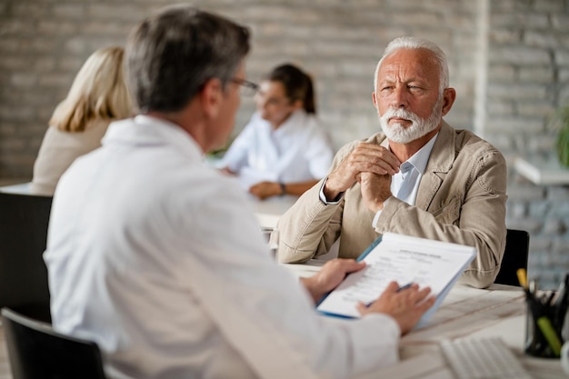 Homme senior et travailleur de la santé communiquant sur les documents d'assurance médicale lors d'une réunion à la clinique