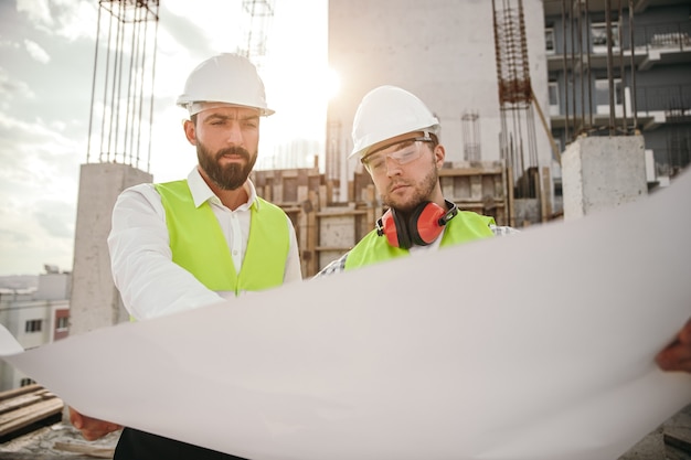 Homme sérieux architecte et ingénieur en casques d'analyse et de discussion de plan tout en travaillant ensemble sur un chantier de construction avec un bâtiment en béton