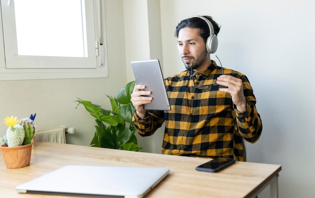 Homme sérieux en chemise à carreaux jaune assis dans un bureau avec une tablette dans les mains et des écouteurs