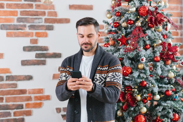 Un Homme Sérieux Et Concentré Correspond Sur Un Téléphone Portable Près De L'arbre De Noël à La Maison