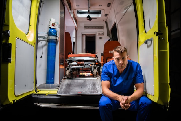 Homme sérieux dans un uniforme médical assis à l'arrière d'une voiture d'ambulance