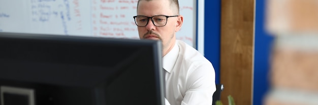 Homme sérieux à lunettes et chemise s'asseoir et regarder l'écran de l'ordinateur. En arrière-plan, un tableau blanc avec du texte.