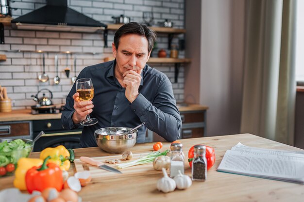 Un homme sérieux se penche à table dans la cuisine et regarde vers le bas. Il tient un verre de vin blanc et la main sur le menton. Légumes colorés et épices avec journal sur table.