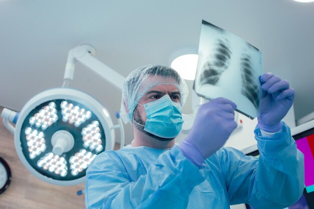 Homme sérieux en uniforme médical protecteur regardant la radiographie pulmonaire