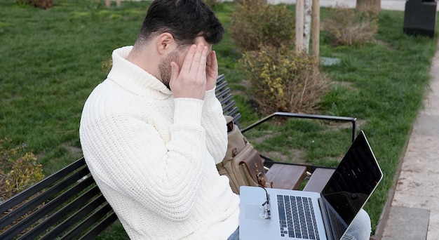 Un homme avec ses mains sur son visage assis sur un banc avec un ordinateur portable.