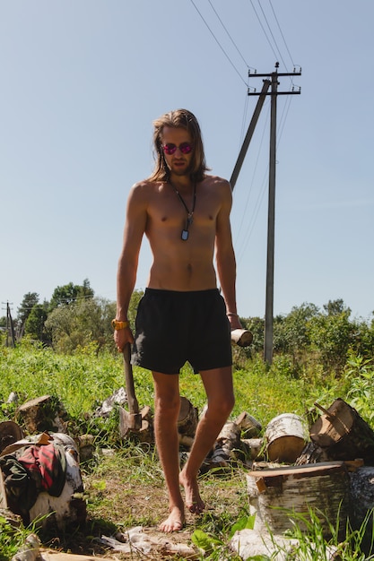 Un homme sexy aux seins nus en short marche avec un hachoir à bois dans une main et une bûche dans une autre entourée d'une corde de bois.