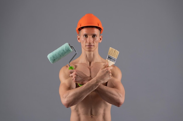 Un homme sexy avec un torse musclé dans un casque tient un rouleau à peinture et un pinceau pour décorer et peindre un fond gris, peintre.