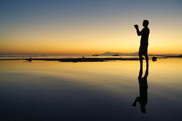 Photo un homme en silhouette photographiant sur la plage contre le ciel au coucher du soleil