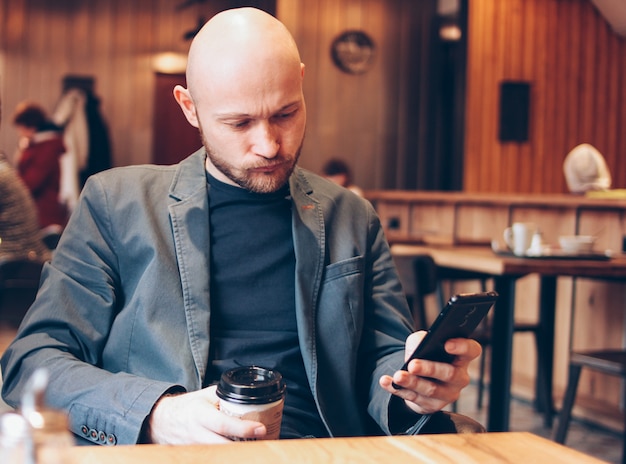 Homme sombre chauve adulte, boire du café dans une tasse en papier et à l&#39;aide de téléphone portable au café