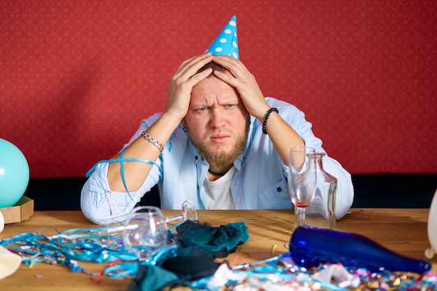 Homme souffrant de la gueule de bois à table avec bonnet bleu dans une pièce en désordre après la fête d'anniversaire