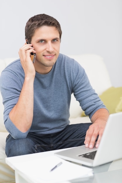 Homme souriant à l&#39;aide de téléphone portable et ordinateur portable dans le salon