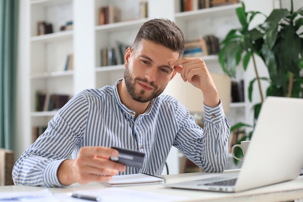 Homme souriant assis au bureau et paie par carte de crédit avec son ordinateur portable.