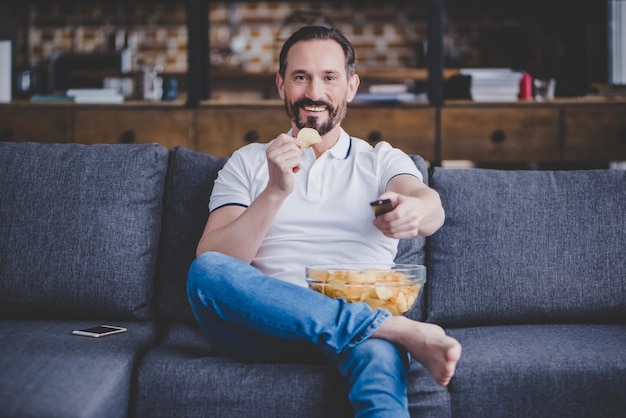 Homme souriant assis sur le canapé devant la télé et mangeant des frites à la maison