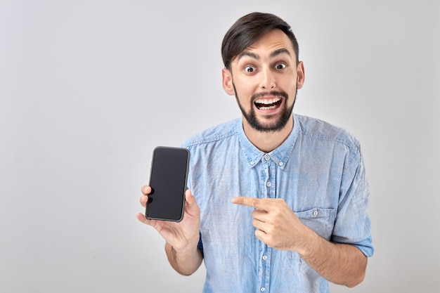 Un homme souriant au visage heureux pointe le doigt sur un téléphone portable à écran blanc isolé sur fond de studio blanc