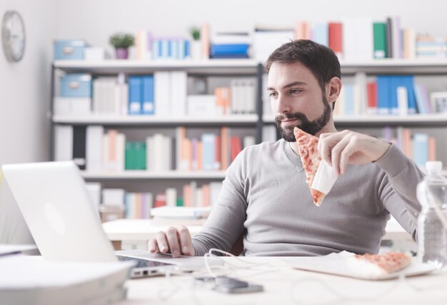 Homme souriant ayant une pause déjeuner au bureau il mange une tranche de pizza et des réseaux sociaux avec un ordinateur portable